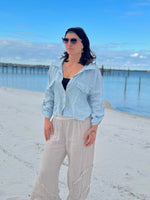 Woman standing on a beach wearing a light blue denim jacket and beige pants.