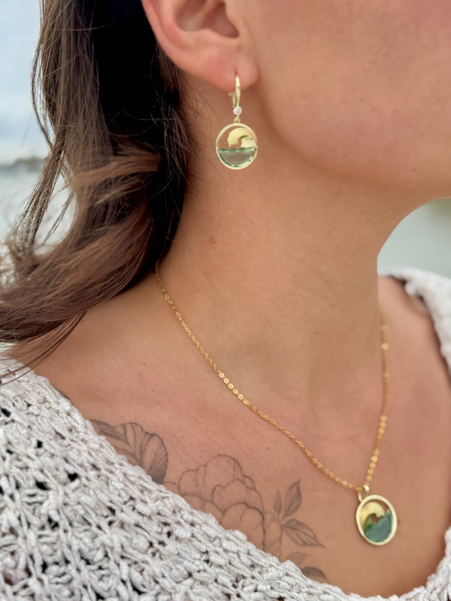 Close-up of a person wearing gold earrings and a necklace with a scenic pendant, outdoors.