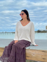Woman sitting by a waterfront wearing a white sweater and purple skirt with sunglasses on.