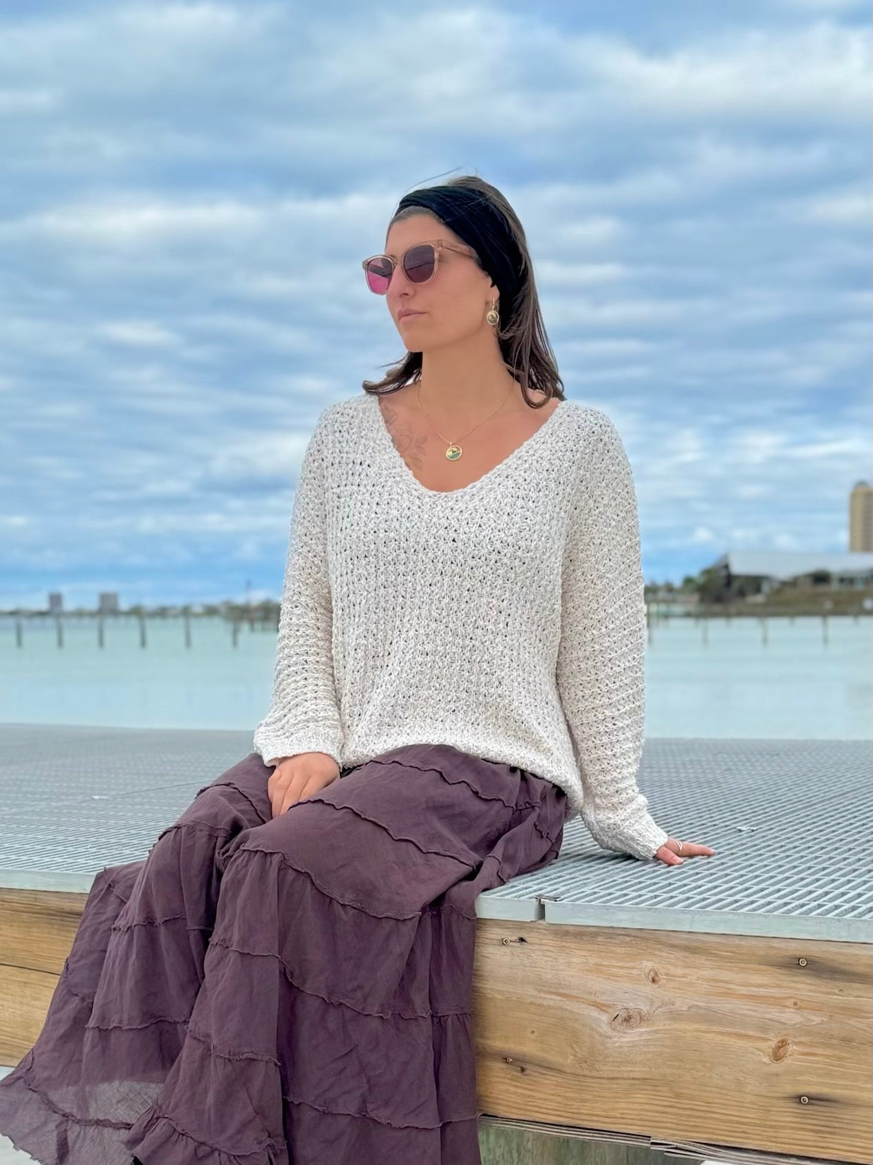 Woman sitting by a waterfront wearing a white sweater and purple skirt with sunglasses on.
