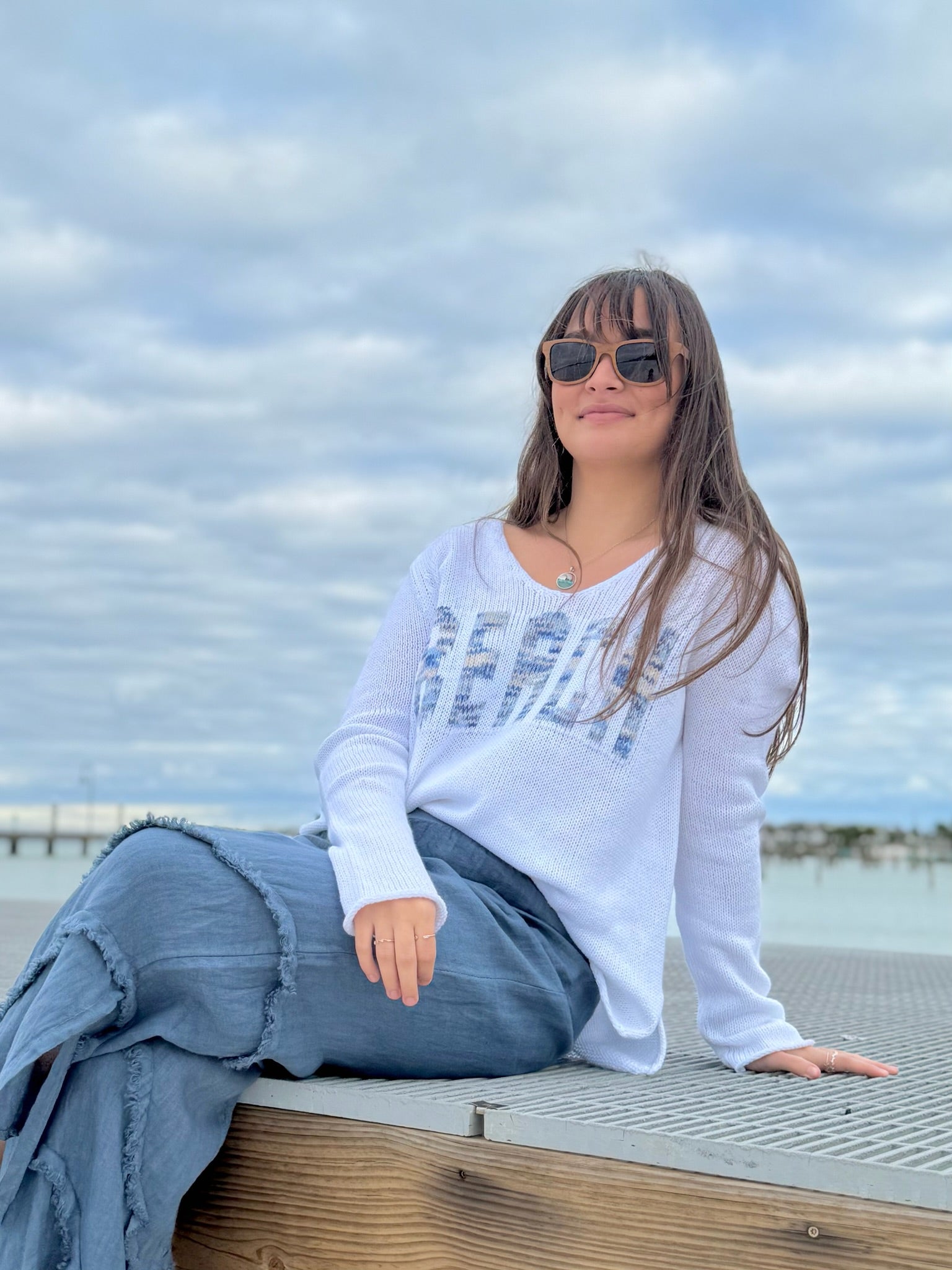 Woman sitting on a dock by the water with sunglasses on, wearing a white sweatshirt with text.