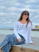 Woman sitting on a dock by the water wearing sunglasses and a white 'Beach' shirt.