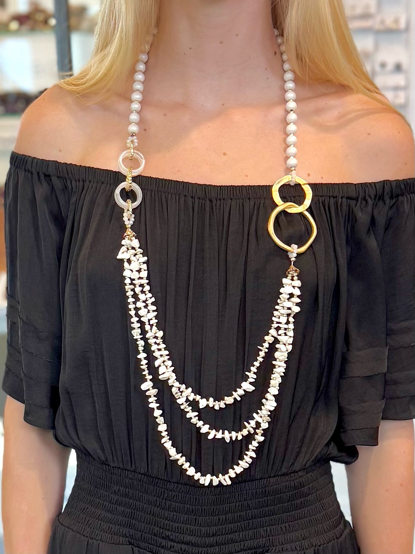 Person wearing a long pearl necklace with gold accents over a black off-shoulder top.