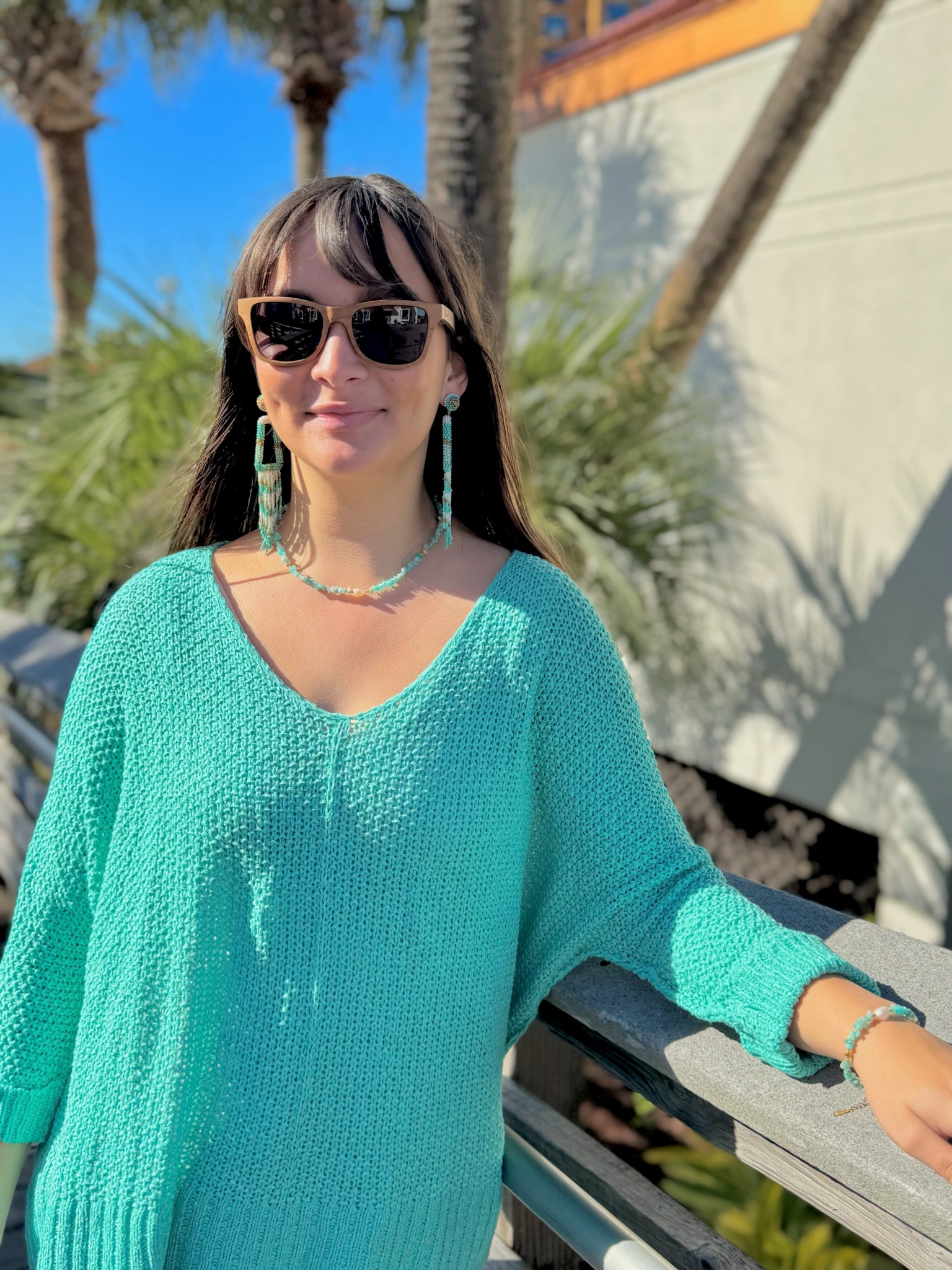 Woman in a teal sweater and sunglasses standing outdoors with palm trees in the background