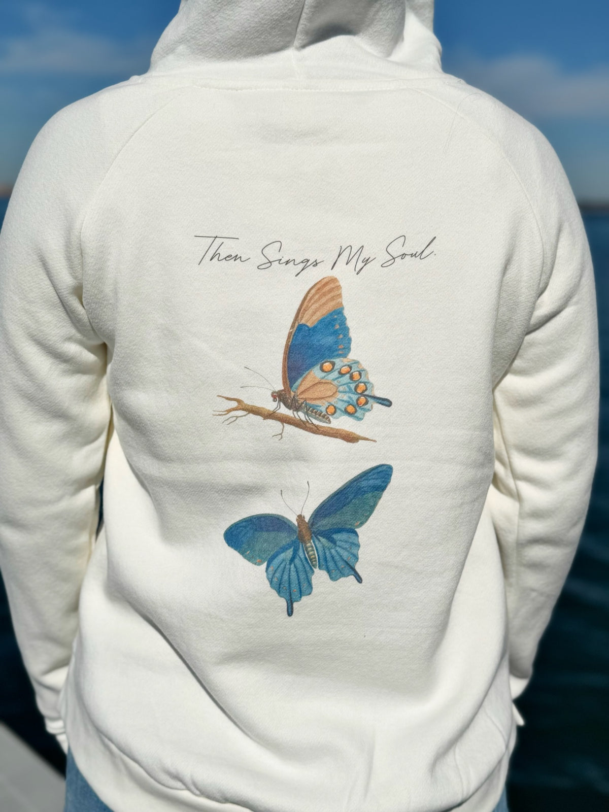 Ivory pullover hoodie with butterfly design on the back against a blue sky.