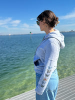 Person wearing a light blue hoodie with butterfly designs by a waterfront