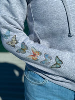 Gray hoodie with watercolor butterflies on the sleeve, worn by a person.