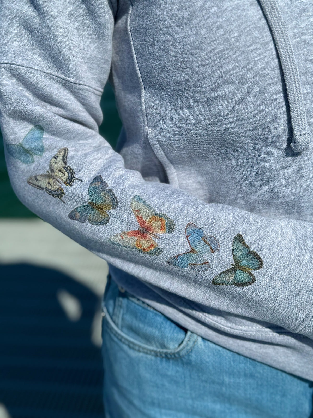 Gray hoodie with watercolor butterflies on the sleeve, worn by a person.