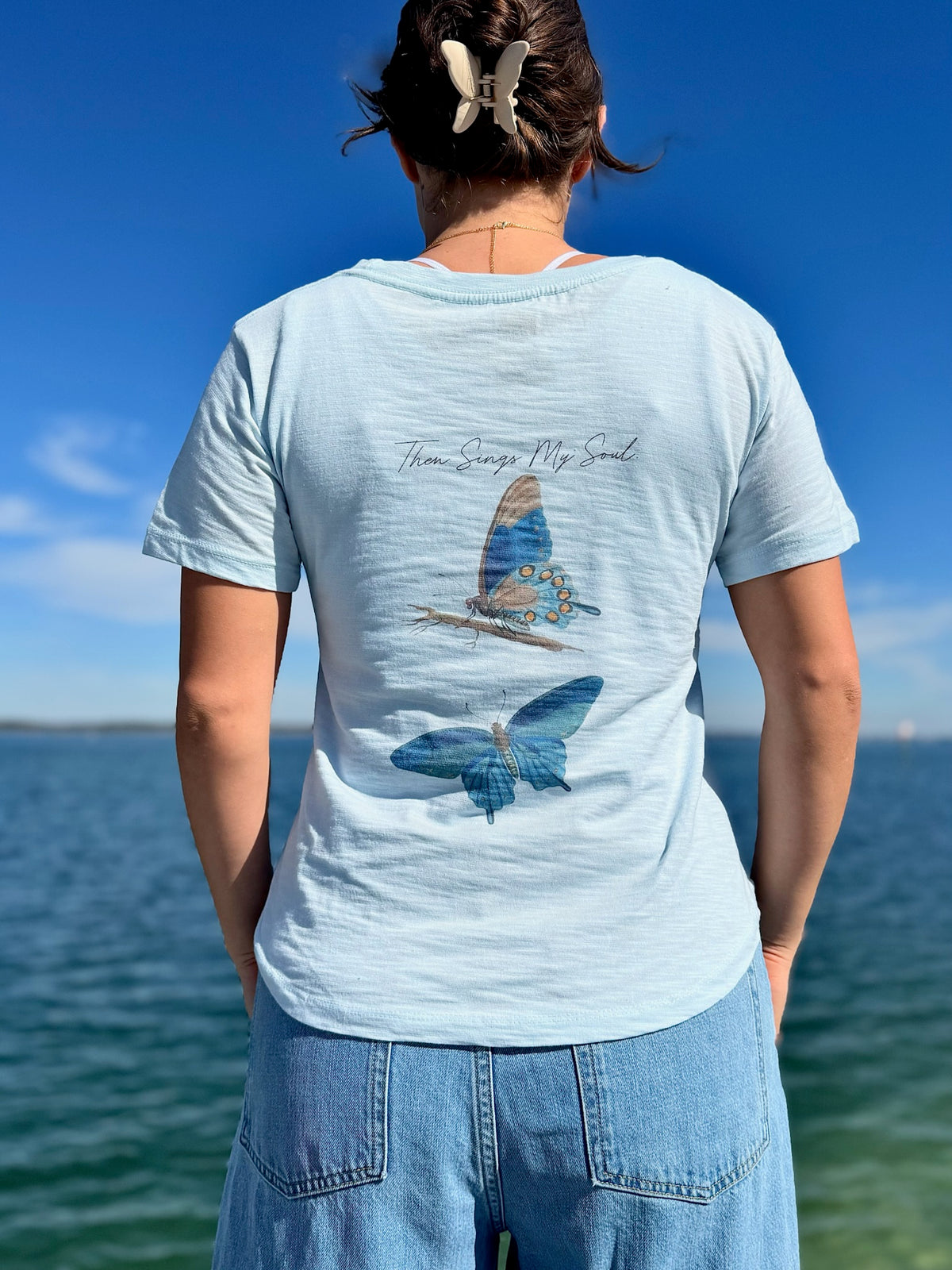 Person wearing a light blue t-shirt with a butterfly design by the ocean