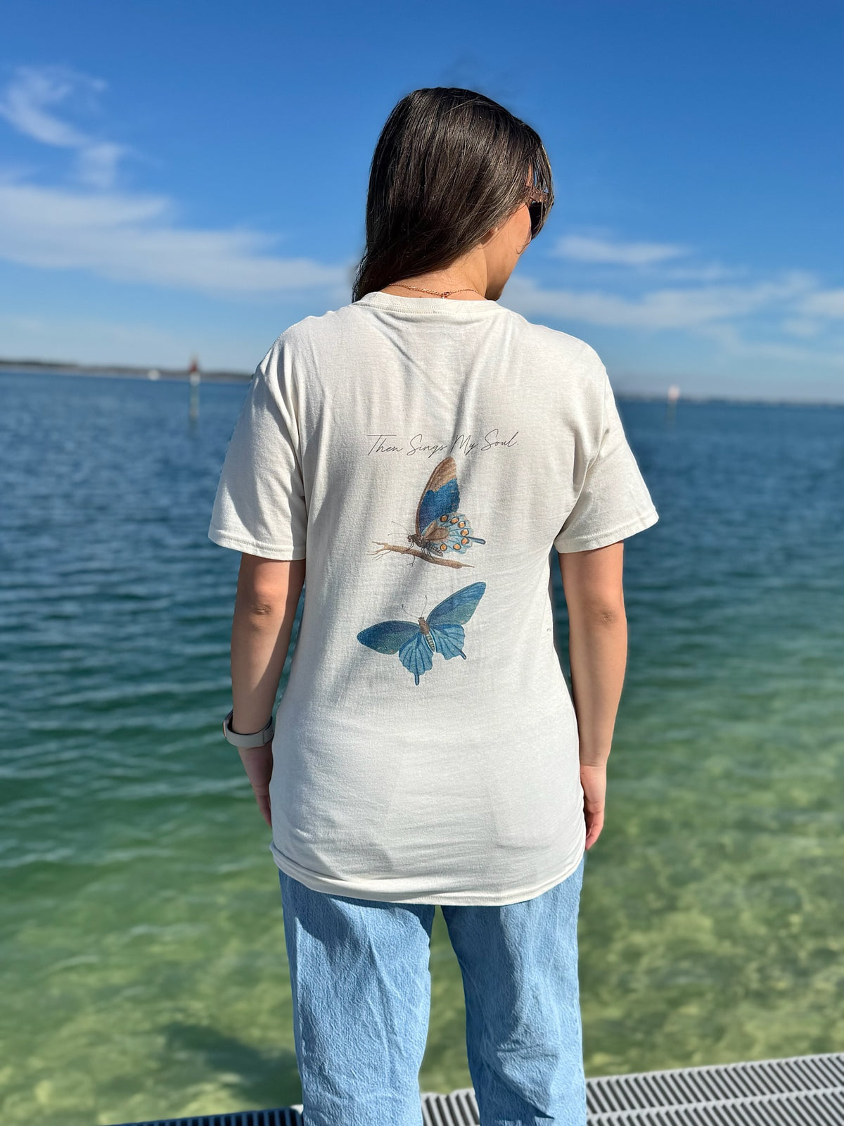 Person wearing a cream t-shirt with a butterfly design by the water