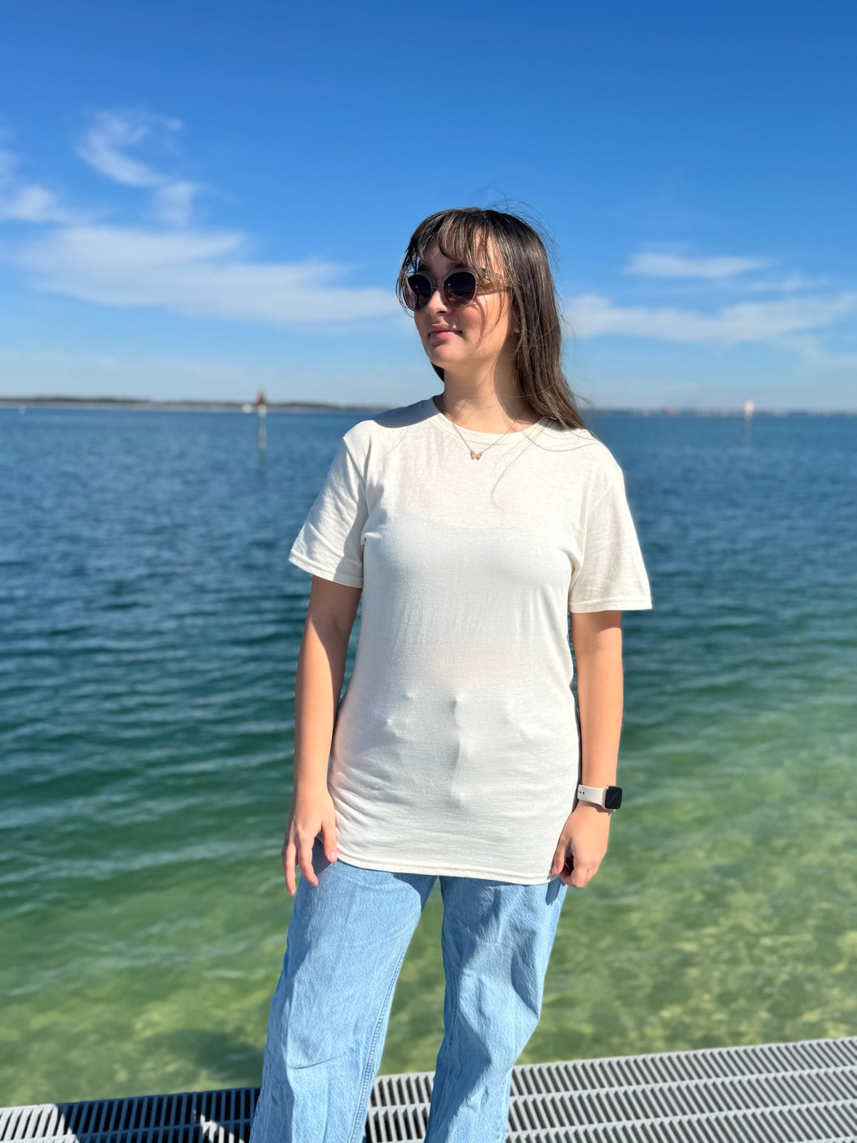 Person standing by a body of water wearing sunglasses and a cream t-shirt.