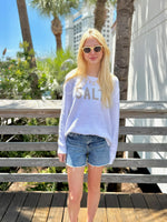 Person wearing a white sweatshirt with 'SALT' text, sunglasses and denim shorts standing outdoors.