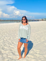 Woman in a 'BEACH' sweatshirt standing on a sandy beach with a clear sky.