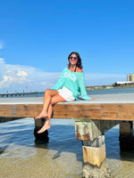 Woman sitting on a dock by the water wearing a 'Beach' sweatshirt.