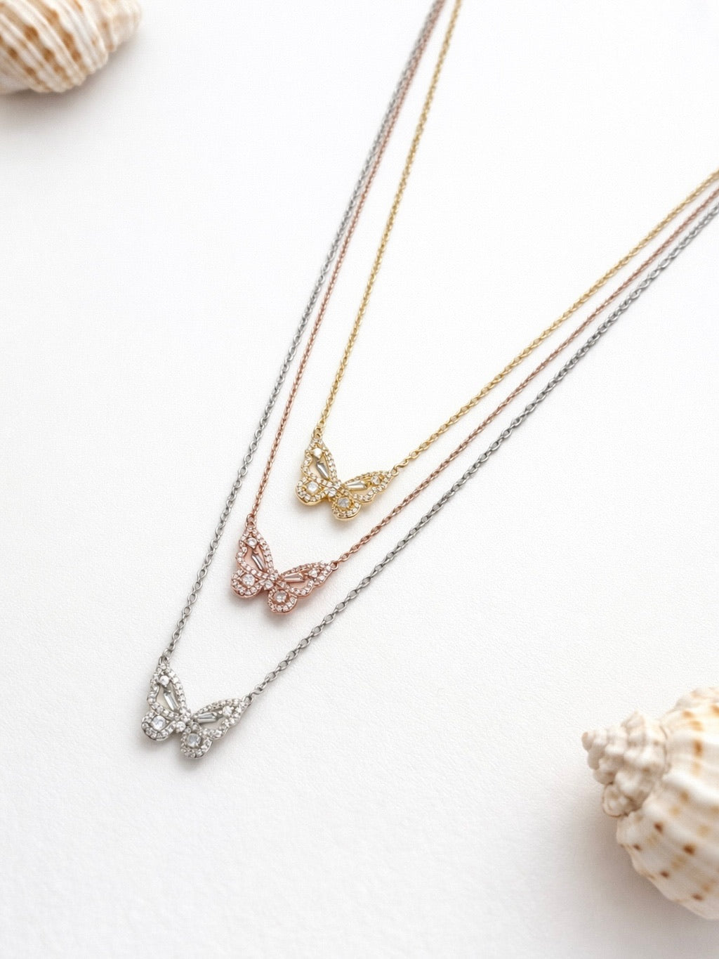Three butterfly necklaces in gold, silver, and rose gold on a white background with seashells.