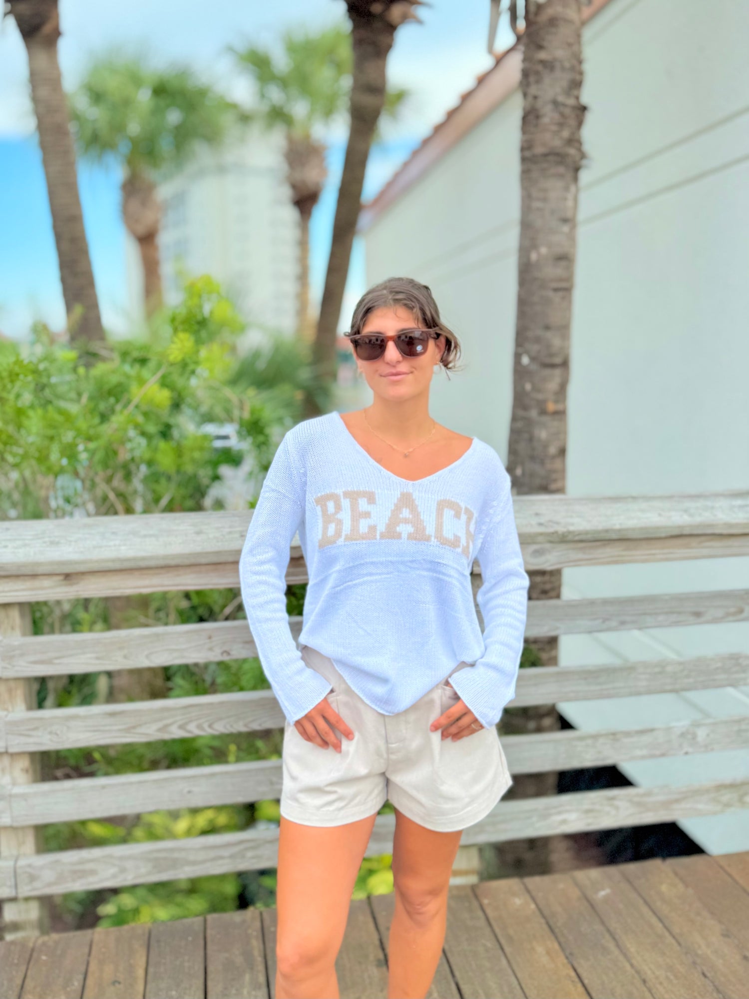 Woman wearing a light blue 'BEACH' sweatshirt and sunglasses, standing on a wooden deck with palm trees in the background.