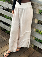 Light beige wide-leg linen pants with frayed seam details and a soft fold-over waistband, styled with a black top and sandals.