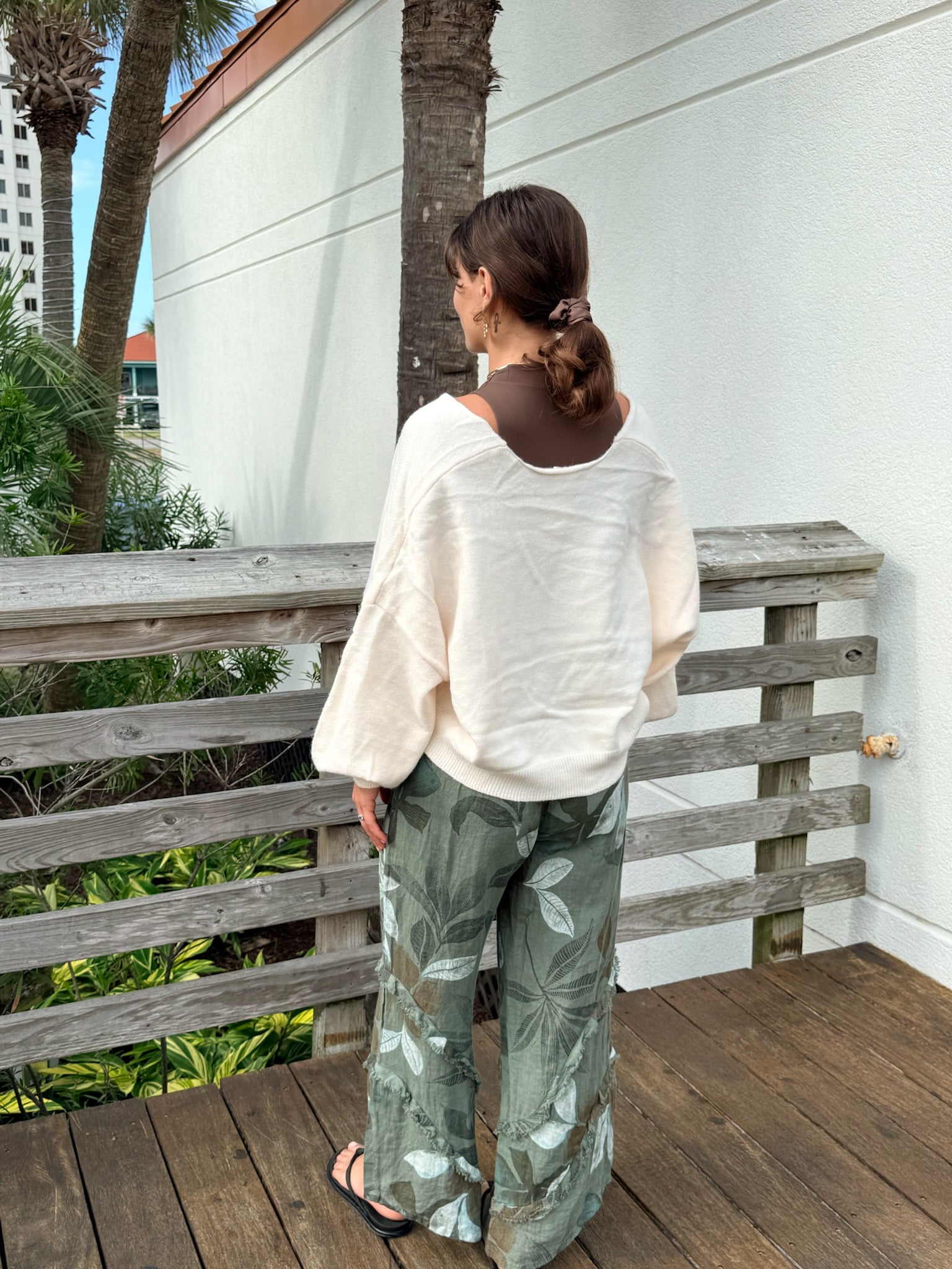 Green floral printed linen pants with wide legs and frayed seam details, styled with an ivory sweater and sandals.