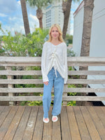 Woman wearing a white sweater and blue jeans standing on a wooden deck with palm trees in the background.