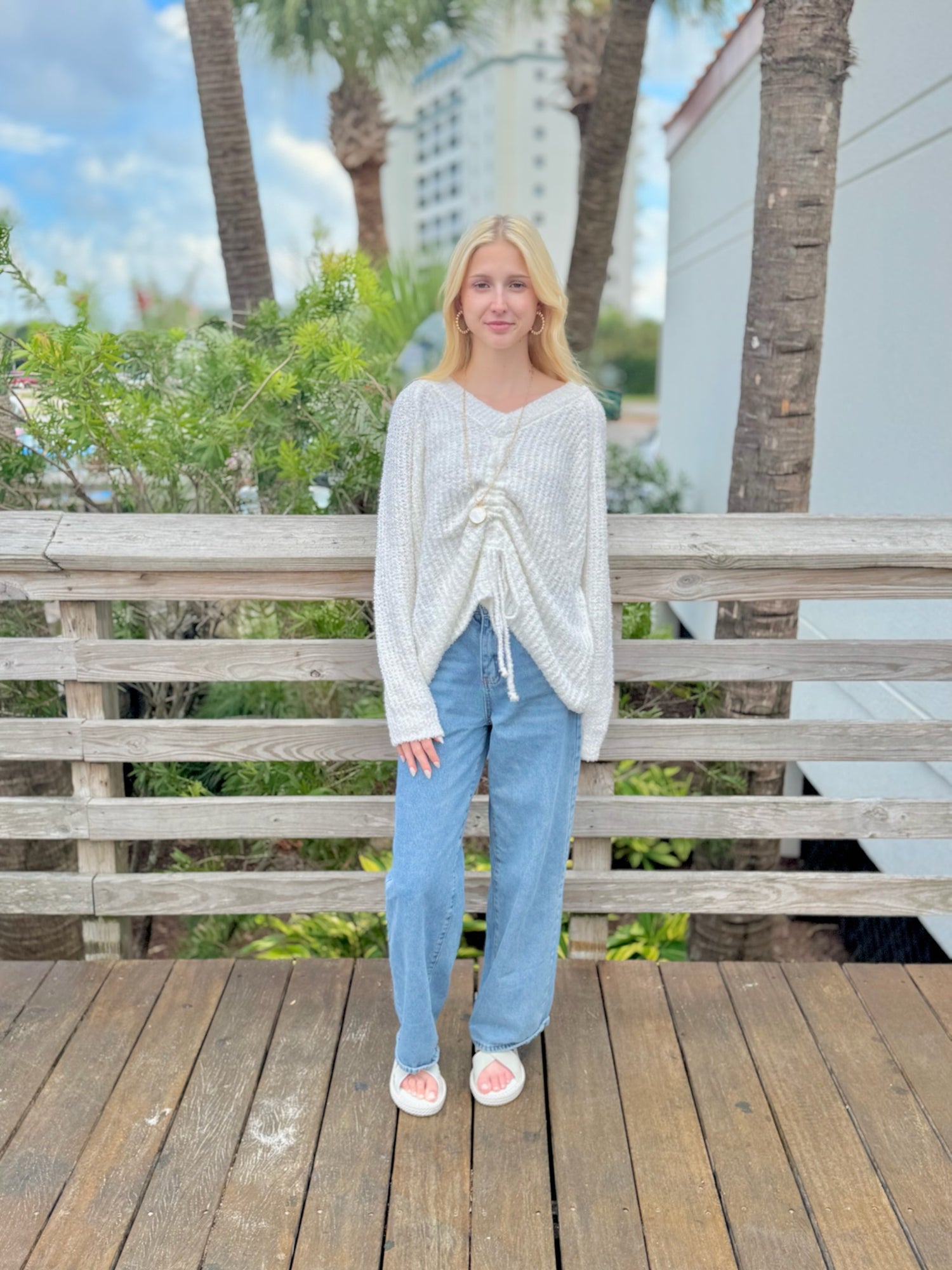 Woman wearing a white sweater and blue jeans standing on a wooden deck with palm trees in the background.
