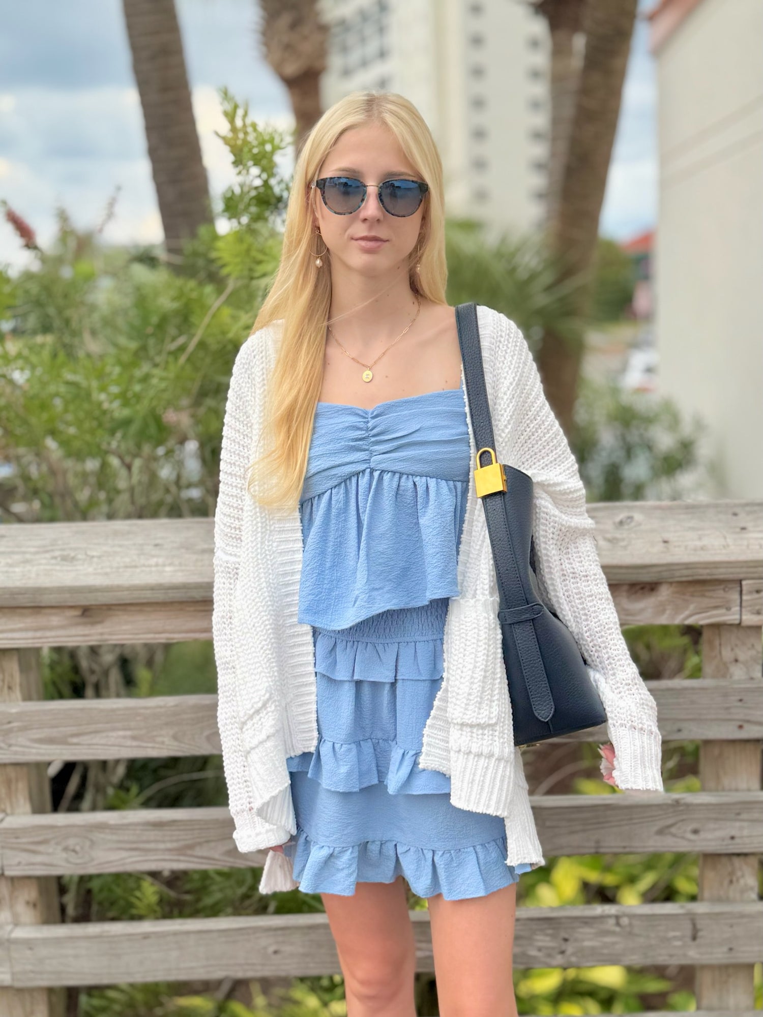 Woman in a blue dress and white cardigan standing on a wooden deck with greenery in the background