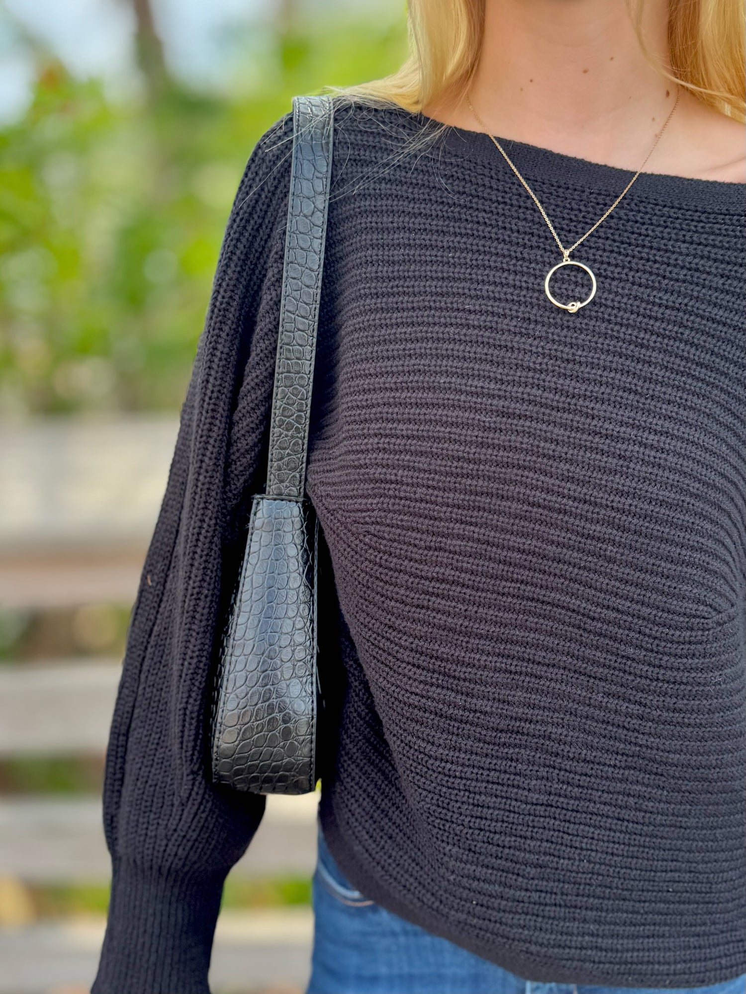 Person wearing a dark sweater with a necklace, carrying a black handbag outdoors.