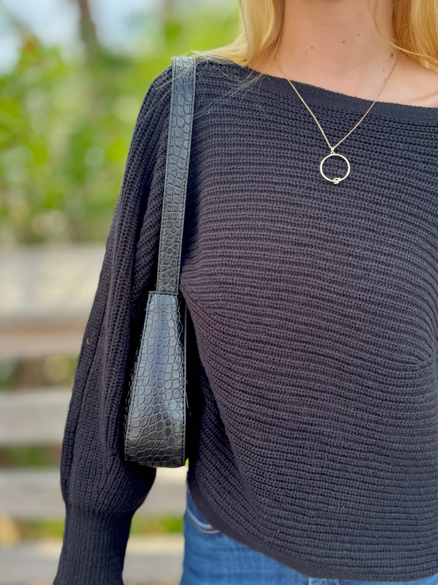 Person wearing a dark sweater with a necklace, carrying a black handbag outdoors.