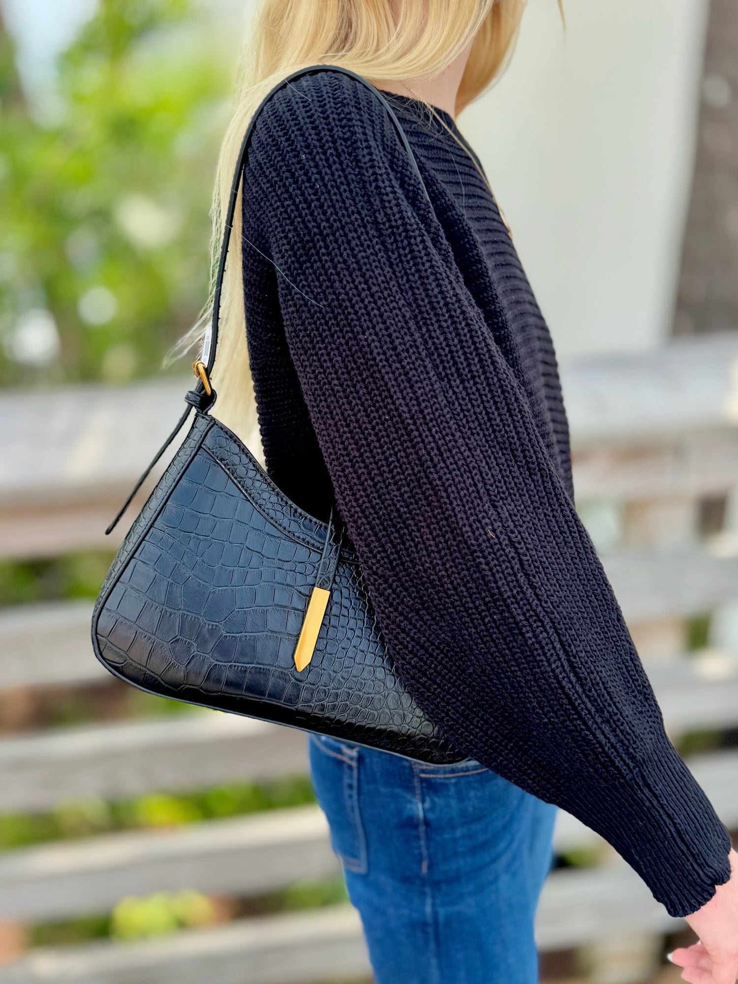 Person wearing a Black  sweater holding a Black handbag with a blurred outdoor background