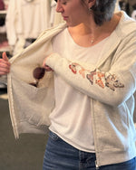 Person wearing a light gray zip-up hoodie with floral embroidery on an arm, holding sunglasses.