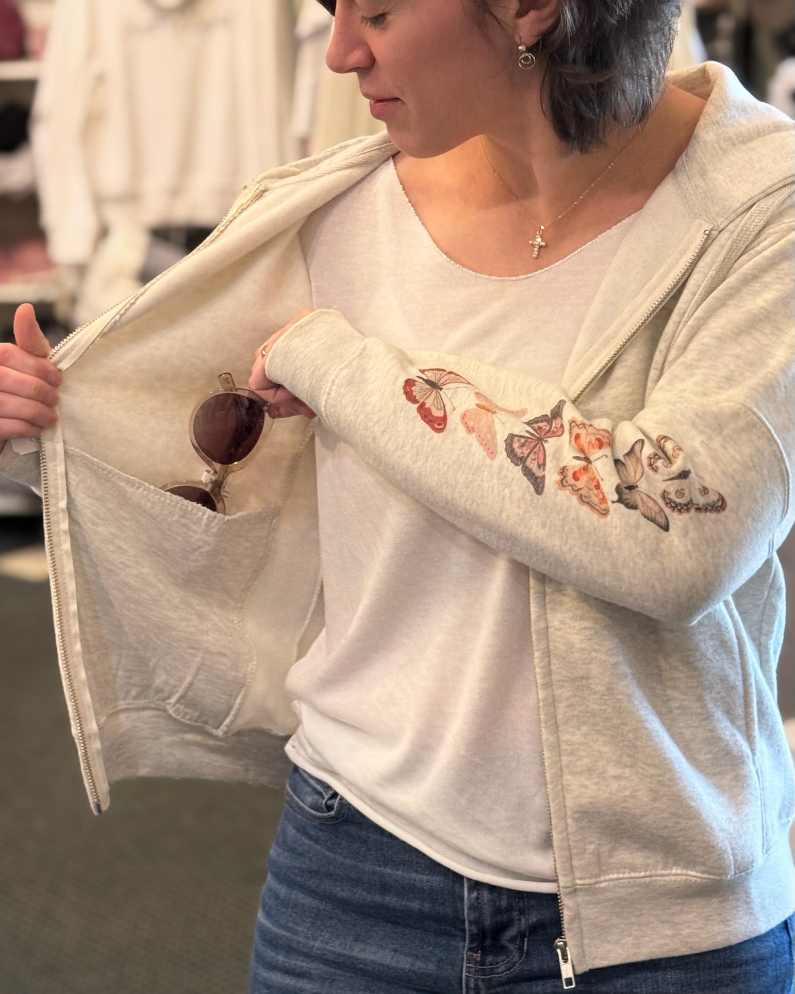Person wearing a light gray zip-up hoodie with floral embroidery on an arm, holding sunglasses.