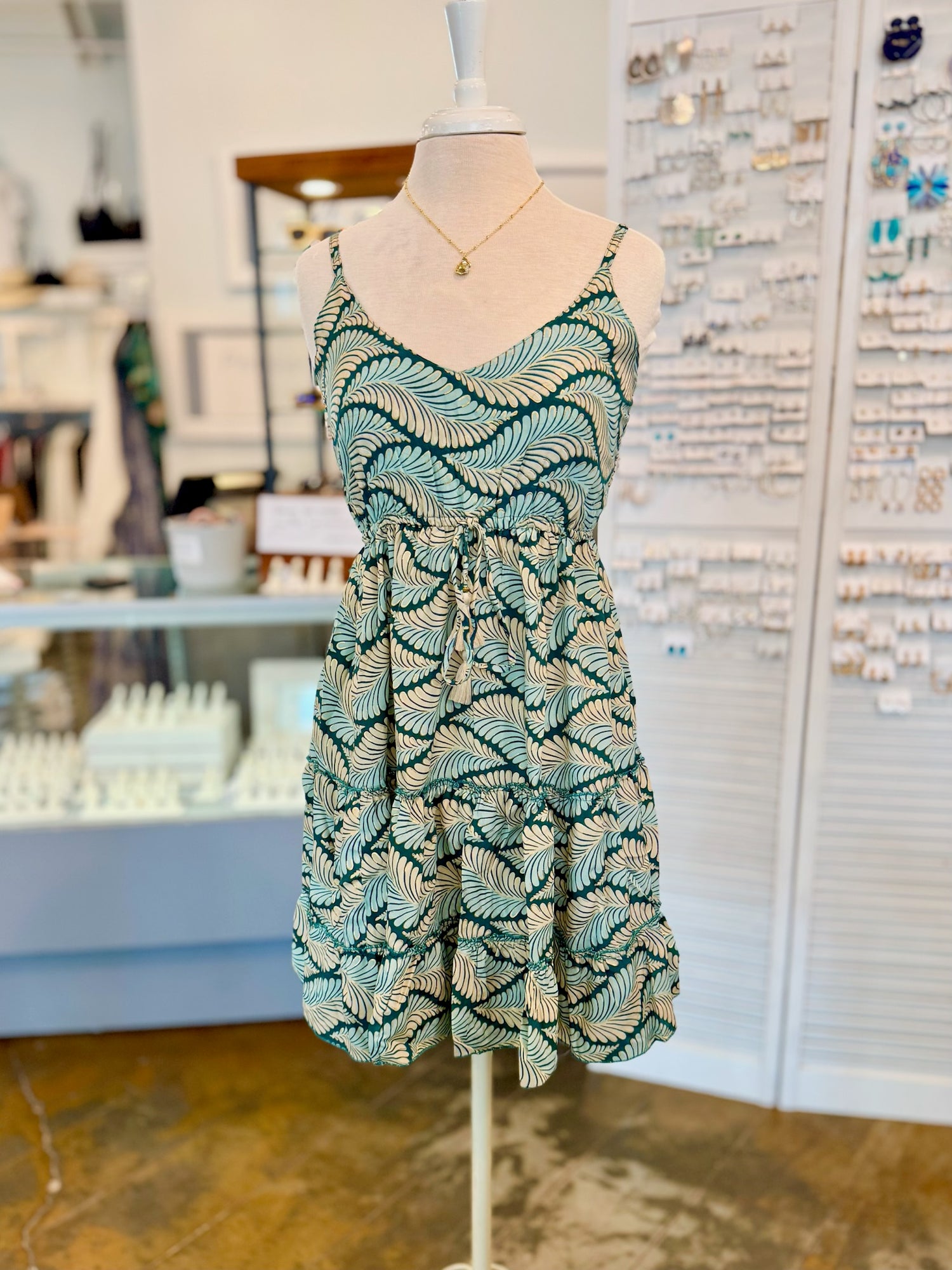 Front view of the Dress with a green and cream leaf pattern, tie waist, and adjustable straps on a mannequin. 