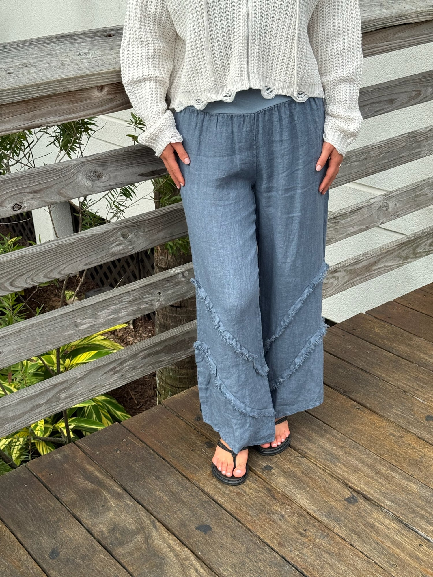 Blue-gray linen wide-leg pants with frayed seam details, paired with a cream knit sweater and black sandals, photographed on a wooden deck.