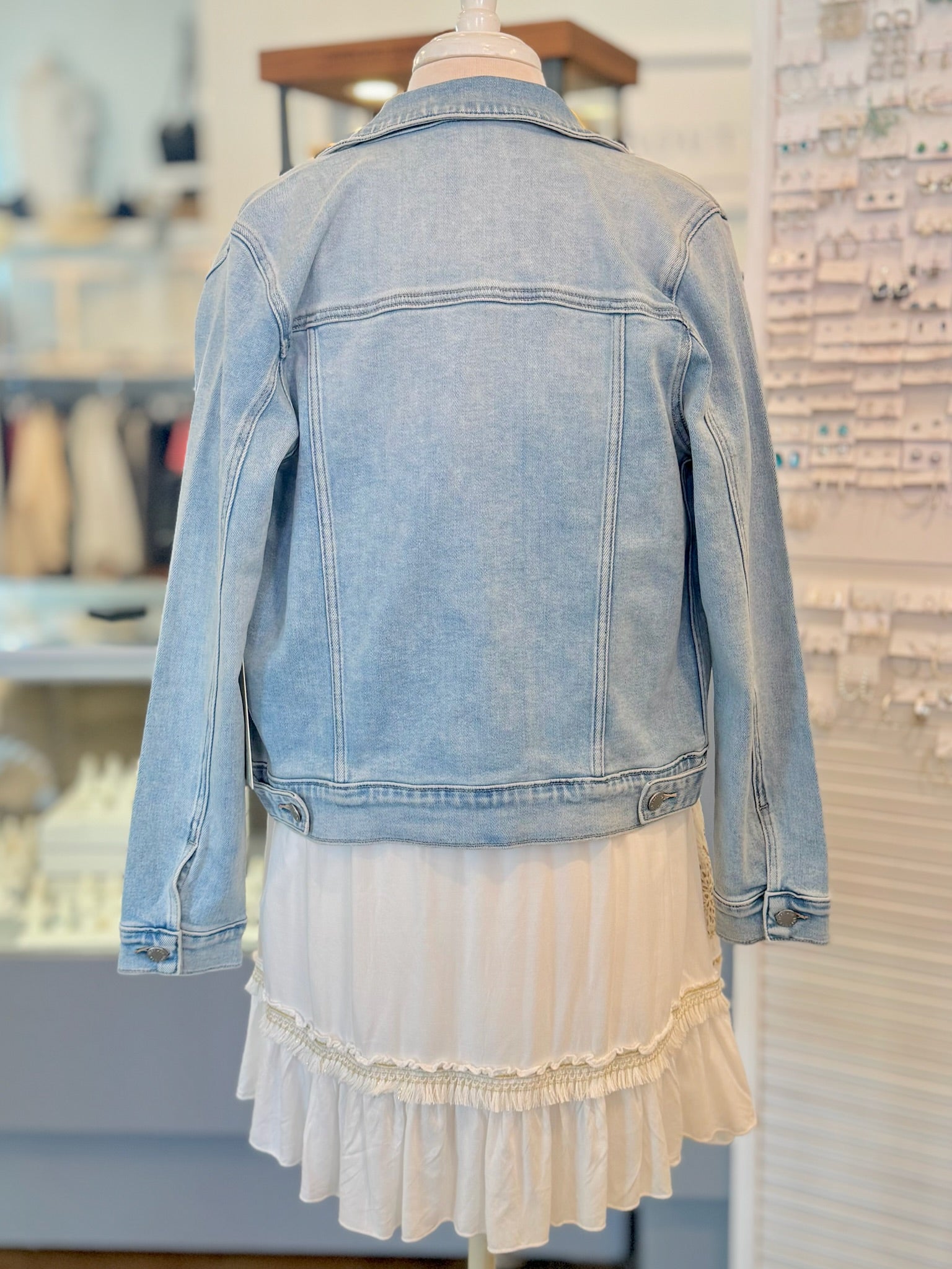 Back view of mannequin wearing a light blue denim jacket over a white dress in a store setting.