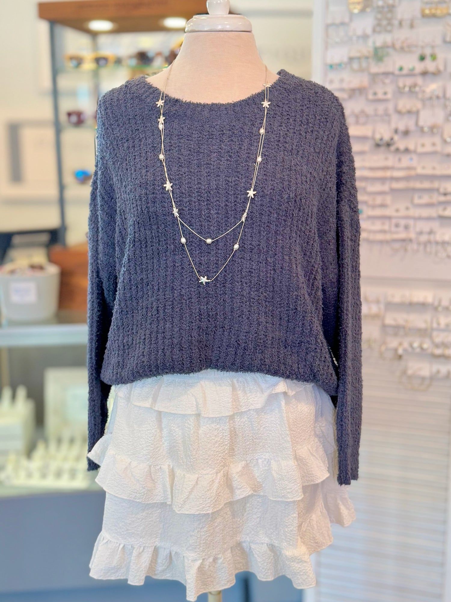 Mannequin wearing a navy sweater and white ruffled skirt with layered necklaces.