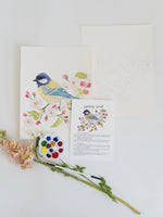 Art print of a bird with flowers on a white surface with paint palette and brush.