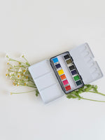 Watercolor paint set with open lid showing colors on a light background with greenery.