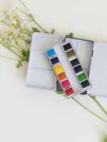 Set of watercolor paints in a compact case with flowers in the background