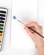 Hand holding a watercolor brush with a small blue stroke on white watercolor paper