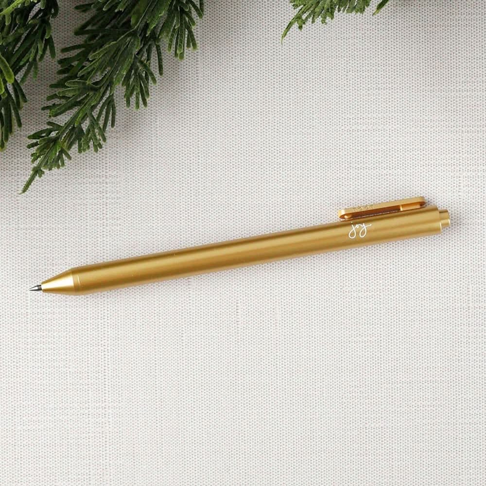 Gold pen with white "joy" text on a light gray fabric background with greenery