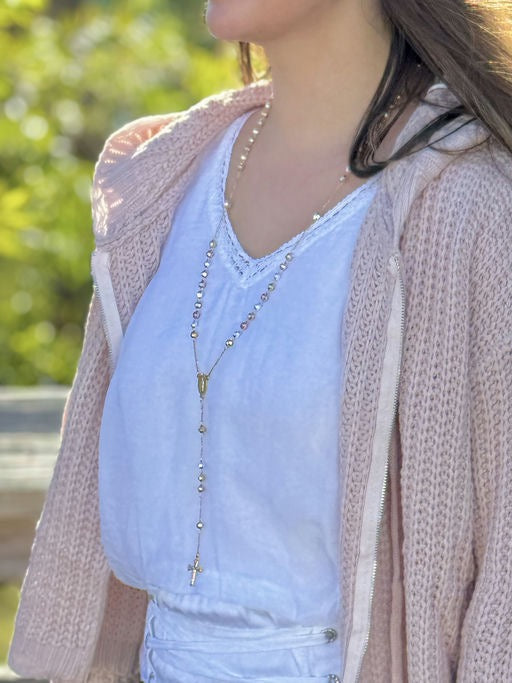 Person wearing a white top and pink jacket with a rosary necklace outdoors.