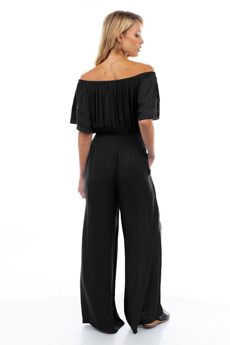A model wearing a black off-the-shoulder jumpsuit with wide-leg pants, featuring a smocked waist and a flowy silhouette.