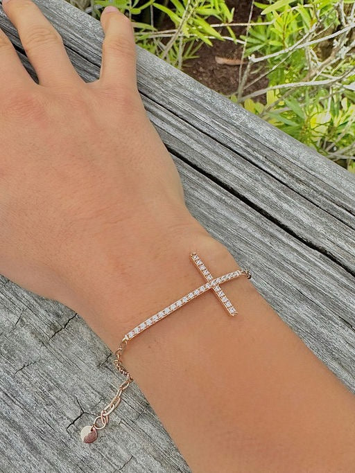 Hand wearing a rose gold bracelet with a clear stone on a wooden surface with greenery in the background