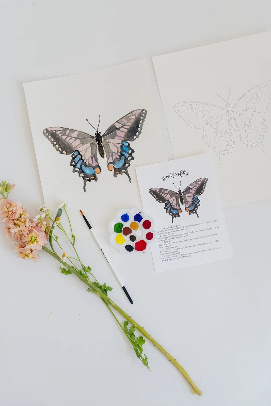 “Butterfly watercolor painting kit with finished butterfly artwork, outline sketch, instruction card, paint palette, brush, and flowers on a white background.”
