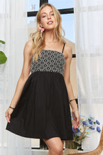 A woman wearing a black babydoll dress with a smocked and embroidered bodice, featuring adjustable spaghetti straps and a flowy silhouette.