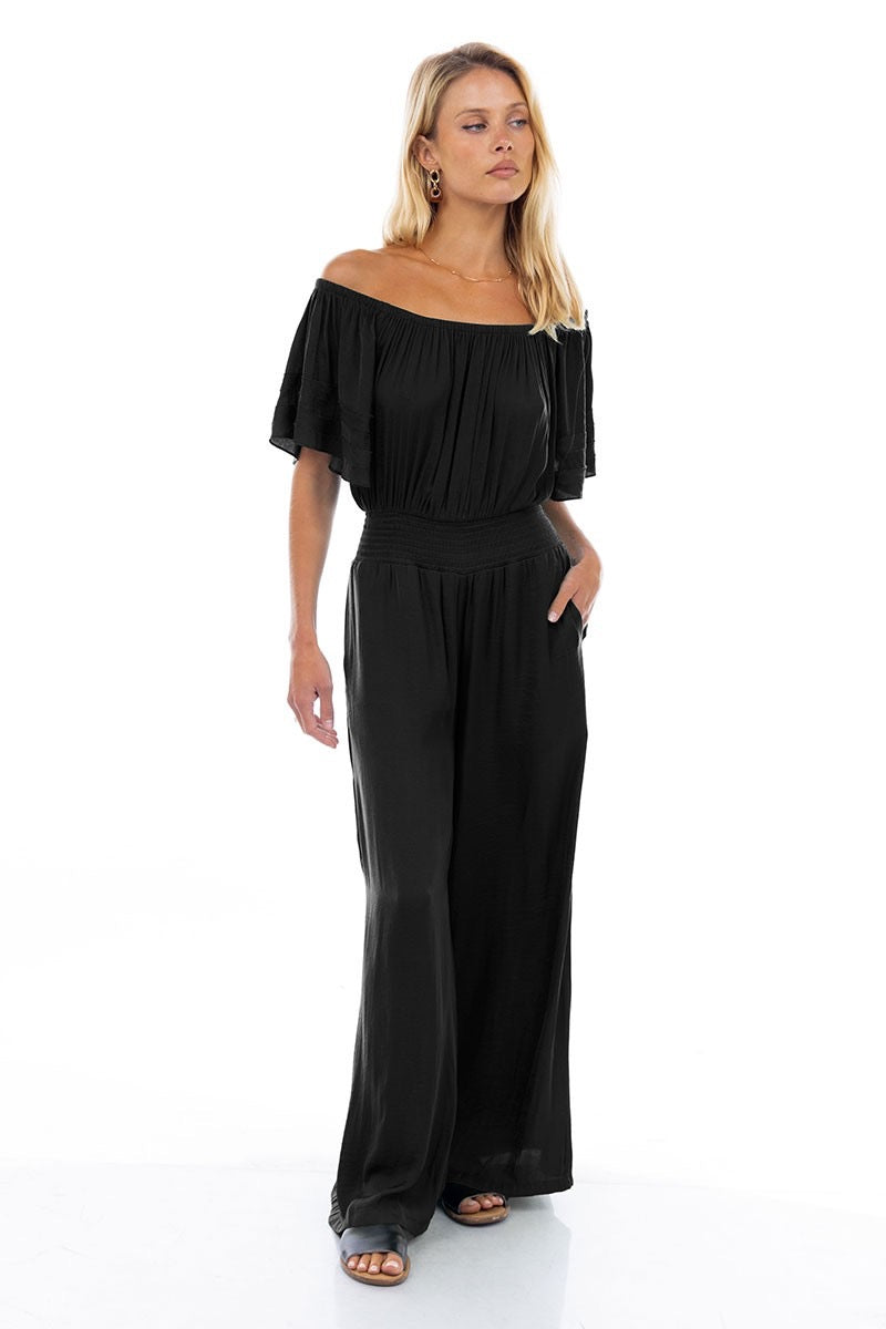A model wearing a black off-the-shoulder jumpsuit with wide-leg pants, featuring a smocked waist and a flowy silhouette.