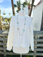 Cable Knit sweater in ivory on a mannequin outdoors with a long bright blue necklace.