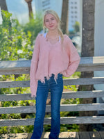 Person wearing a pink sweater and blue jeans standing on a wooden deck with greenery and buildings in the background.