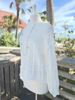 Side view of Cable Knit sweater in ivory on a mannequin outdoors with a long bright blue necklace.
