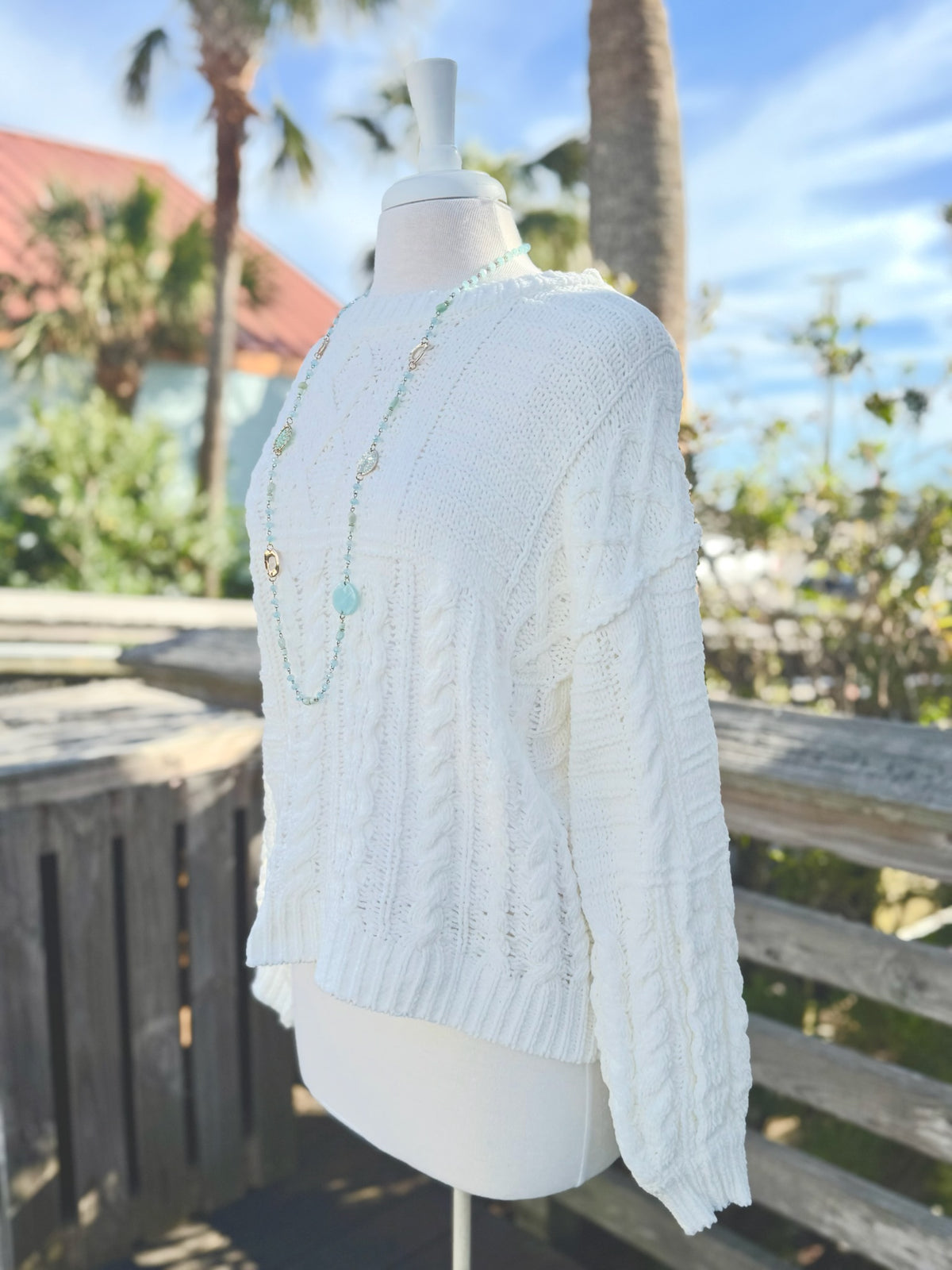 Side view of Cable Knit sweater in ivory on a mannequin outdoors with a long bright blue necklace.