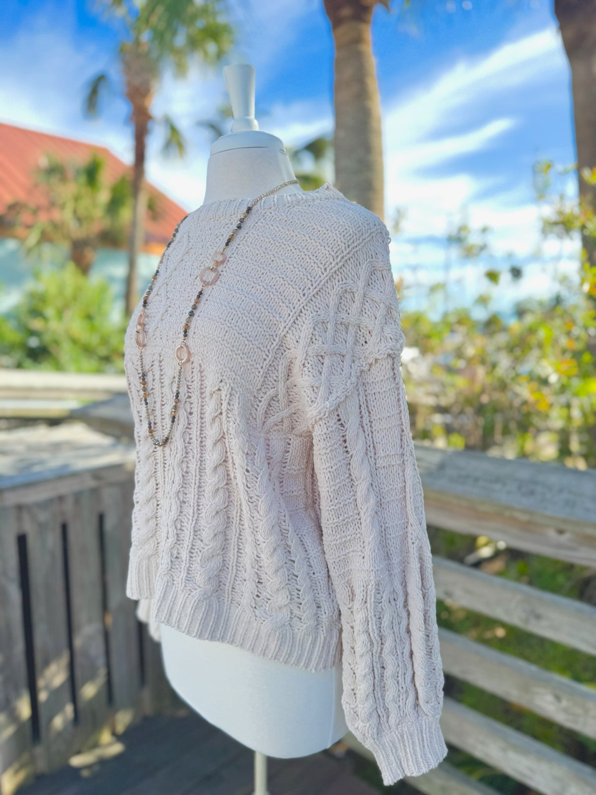 Cable knit sweater in beige on a mannequin outdoors with a long taupe necklace
